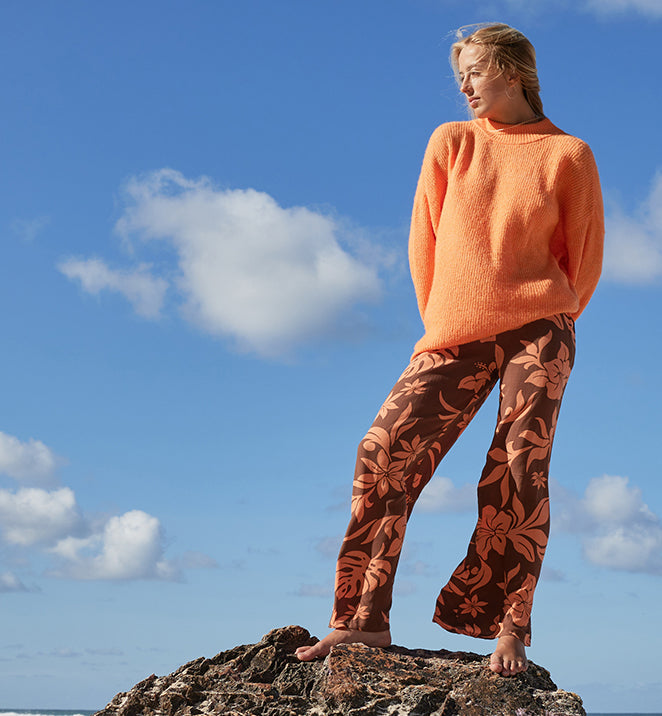 A woman in an orange sweater and floral pants stands on a rock, enjoying the scenery