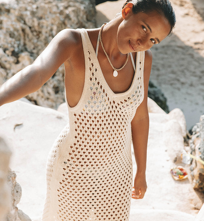 Smiling woman in a knitted beach dress stands on stone steps near a sandy beach with a rocky background conveying a summer vibe.