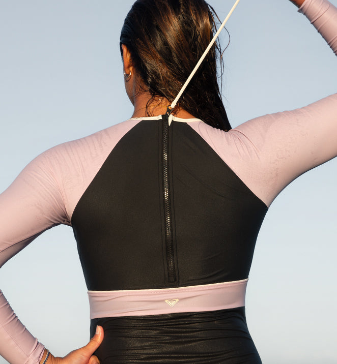 A person wearing a summer rash shirt with light pink sleeves and a black back is pulling a zipper cord. The image conveys a sense of readiness against a clear sky.